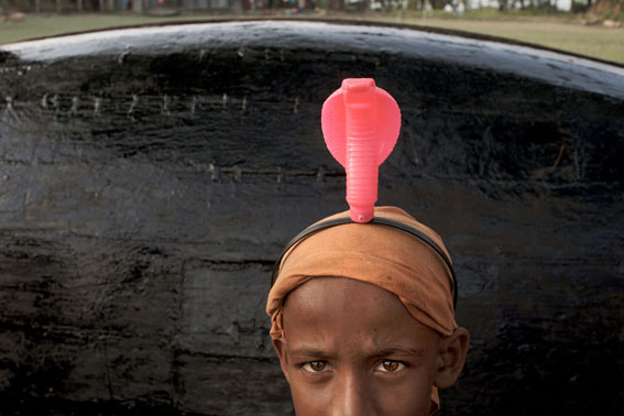 Climate change, Bangladesh, 9.2009. A young boy in Forki village proudly displays his plastic cobra 'hat', his back against the tarred bottom of a fishing boat.